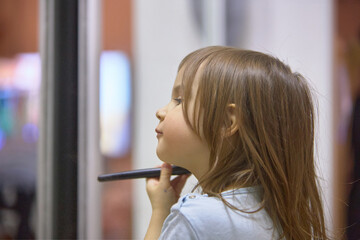 A little girl holds a blush brush to apply powder on her cheeks and face, happily looking in the mirror. Cute baby is playing makeup makeup game..