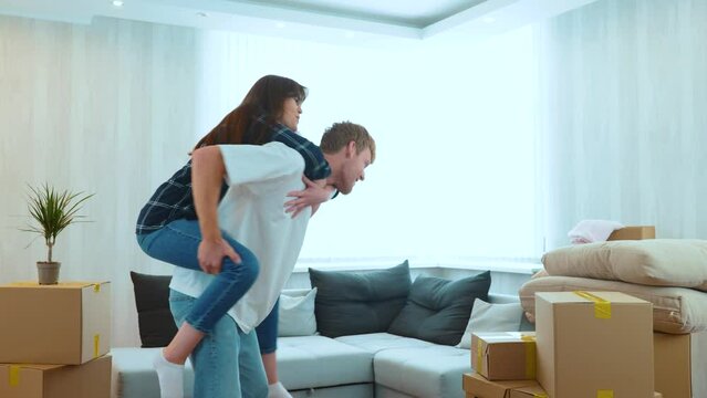 Close up of cheerful Caucasian man and woman having fun playing in new house on moving day. Piggyback ride. Happy young couple in room with many boxes moving in together. Home concept. Real time