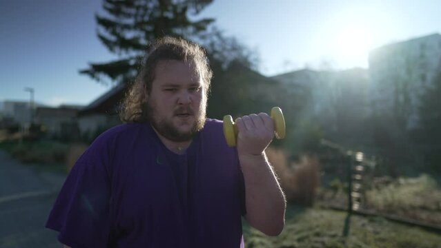 One young overweight man working out lifting dumbbells outside. A fat chubby guy training outdoors