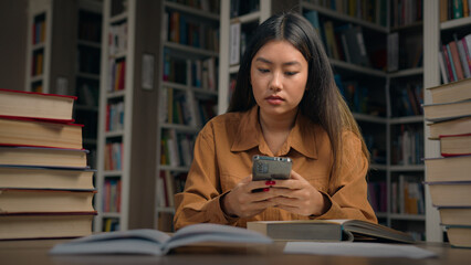 Young serious asian woman student sitting in library at desk searching information on Internet...