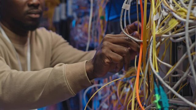 Cropped shot of Black IT engineer connecting tangled server wires in operational rack server cabinet of data center
