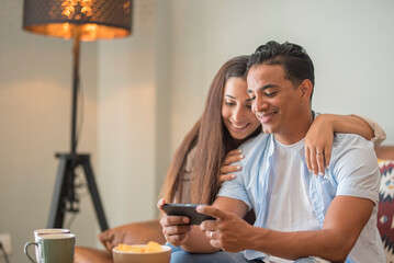 Smiling millennial couple sit on sofa in living room have fun using modern smartphone devices together, happy young husband and wife laugh relax at home browsing application on cellphone gadgets web