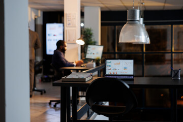 Laptop computer with company investment graph on screen, standing on desk in startup office. In background businessman analyzing marketing statistics, typing strategy. Business concept