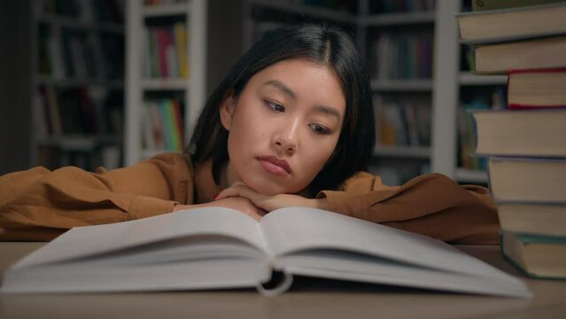 Bored sad tired lazy young korean woman read boring book lying on desk in university library unmotivated female student doing homework preparing for college exam overload with study unloving subject