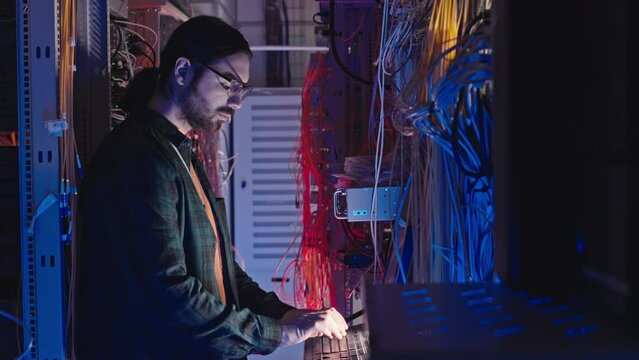 Side view medium shot of Caucasian male IT administrator working on laptop while standing in dark server room of data center