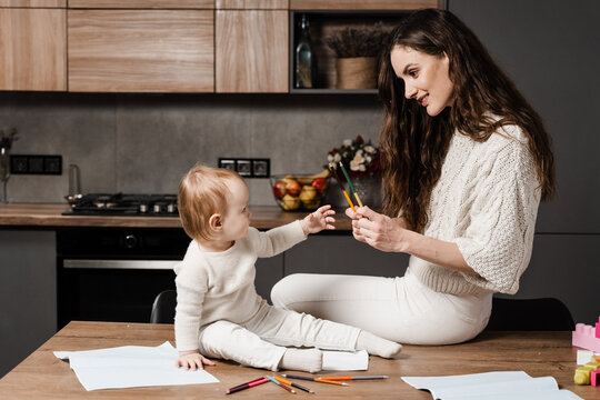 Mom and daughter toddler fool around and play together in the kitchen at home. Happy family of mom and daughter laugh and play with colored pencils while drawing. Maternity leave.