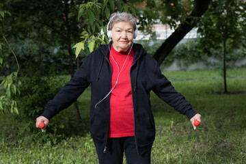 Smiling elderly woman is doing exercises with dumbbells outdoors in the yard.  Active life of pensioners. Adaptation of pensioners in the modern world. Prevention of Alzheimer's disease, sclerosis etc