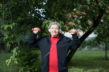 Smiling elderly woman is doing exercises with dumbbells outdoors in the yard.  Active life of pensioners. Adaptation of pensioners in the modern world. Prevention of Alzheimer's disease, sclerosis etc