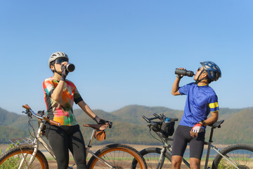 Fototapeta na wymiar Tired Asian cyclist couple taking a break to drink water from a bottle with a beautiful lake and mountains in the background.
