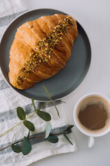 Flat lay of cup of cappuccino with kitchen towel, croissant and eucalyptus branch on the table