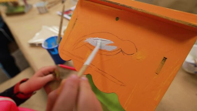 Hands of the child in pink jacket and red watch draw with brush and paints a sketch of a bird with gray paint on the bright orange roof of the plywood craft. Painting a homemade birdhouse