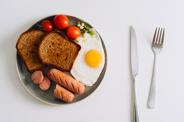 Breakfast plate with knife and fork on the white table, grilled sausage and whole wheat toast with fried egg and cherry tomatoes on a plate