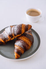 Close up two chocolate croissants on a plate with cup of cappuccino in the background