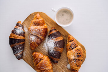 Cup of coffee and a bunch of appetizing brown and chocolate croissants with powdered sugar on a wooden board on white table, flat lay