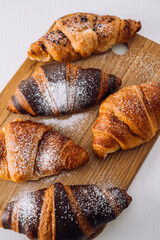 Close up bunch of appetizing brown and chocolate croissants with powdered sugar on a wooden board on white table