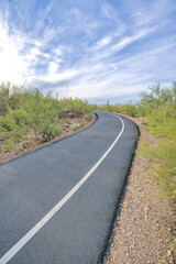 Fototapeta na wymiar Uphill asphalt bike path and walking pathway at Tucson, Arizona. Pathway with white line on the right in the middle of desert land and slope against the sky background.