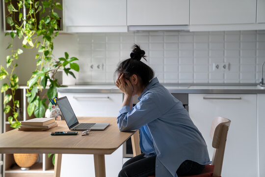 Unhappy frustrated young woman sitting at kitchen table looking at laptop screen having problems in job searching, reading bad news via email. Upset student girl feeling unmotivated to study online