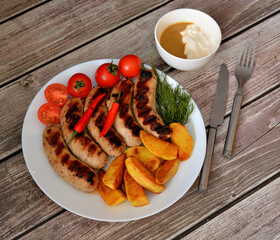 A plate with grilled sausages, cherry tomatoes, herbs, fried potatoes with hot peppers and a bowl with sauce on a wooden table.