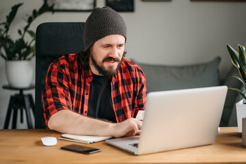 Successful clever caucasian young man, student, freelancer, programmer, sitting at home in kitchen, taking notes in a notebook during an online webinar or lecture, working on a project, using laptop