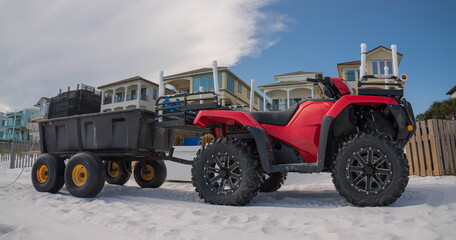 ATV with trailer on white sand shore of Destin, Florida beach. Red ATV with black trailer against the beach houses and giant cloud in the sky background.