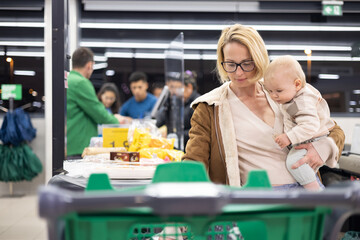 Mother shopping with her infant baby boy, holding the child while stacking products at the cash...