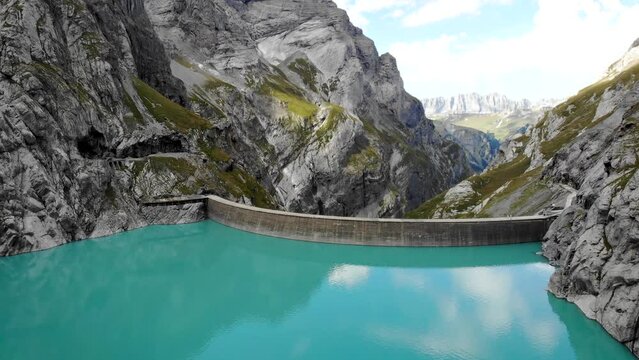 Aerial flyover over turquoise waters of lake Limmernsee and hydroelectric dam in Linthal Glarus, Switzerland with cliffs and mountain peaks in background