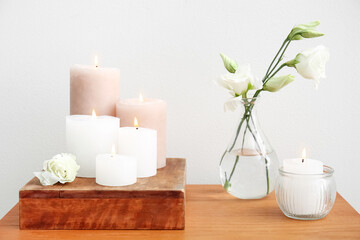 Burning candles and vase with flowers on table near white wall
