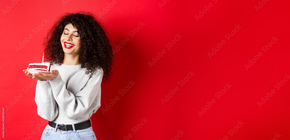 Wall mural Happy birthday girl celebrating and making wish, holding bday cake and smiling, standing on red background - Wall murals