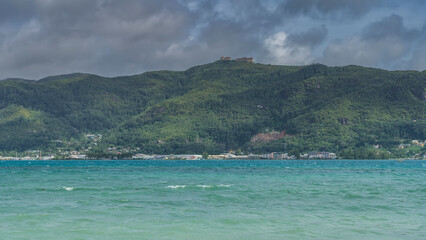 Obraz premium The hilly slopes of the tropical island are overgrown with green vegetation. At the top of the ridge, against the sky and clouds- the building. Ripples on the surface of the turquoise ocean.