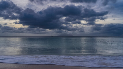 Obraz premium Evening seascape in shades of blue. Picturesque clouds in the sky. Yachts and the silhouette of the island on the horizon. Glare on the ocean surface. Foam of waves on a sandy beach. Long exposure