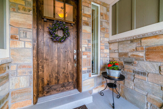 Utah- Dark wood front door with window panel above the lavender wreath and sidelight panel. Entrance of a house with stone veneer siding and wood front door with potted flowers on the right.