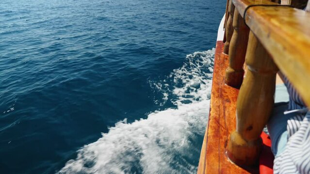 Side view of a vessel (boat, boat, ship) sailing in the Mediterranean Sea. Wooden railing against the background of the sea. Deep blue sea, vessel splits shiny frothy white waves. Sea stroll