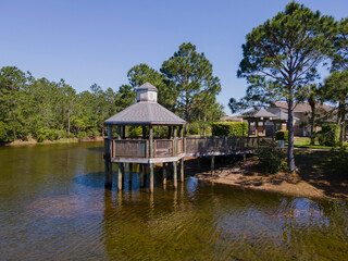 Gazebo over shallow pond at a residential neighborhood in Navarre Florida. The recreation area has a relaxing view of water reflecting the lush green trees and blue sky.