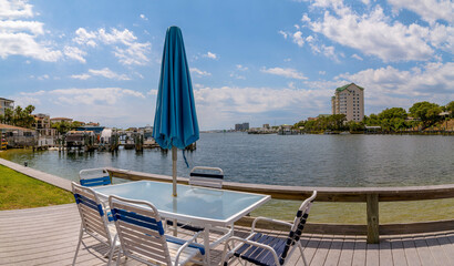 Outdoor dining table with chairs and closed umbrellas on a deck at Destin Harbor, Destin, Florida. Outdoor lounge with views of boats on water and buildings against the bright sky with clouds.