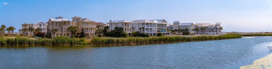 Fototapeta na wymiar Lakefront homes on the beach at Destin Point, Destin, Florida panorama. There is a lake at the front with tall grasses on the shore near the three-storey houses against the bright sky background.