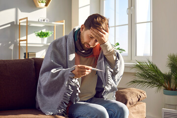 Sick young man sitting on sofa and checking his temperature wrapped in blanket and touching his...