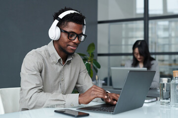 Young African American businessman in wireless headphones typing on laptop during his work at table in office - obrazy, fototapety, plakaty