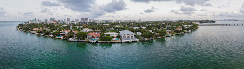 Panorama of Miami Beach Florida with buildings surrounded by calm water. Aerial coastal landscape of waterfront houses against cloudy blue sky.