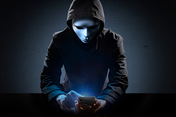 仮面を被り、スマートフォンを操作している犯罪者。
ハッカー、ネット中傷などのイメージ
