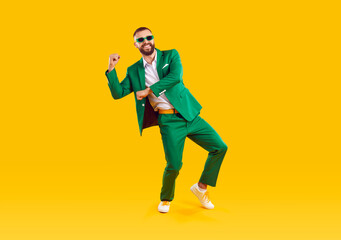 Happy carefree handsome young man in trendy green suit and sunglasses celebrating St Patrick's Day, having fun and dancing on yellow colour studio background. St Patrick's Day and fashion concept