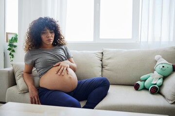 Pregnant woman sitting on the couch lower back pain and headache, strain on the spine during pregnancy. Lifestyle difficulties of motherhood preparation for childbirth, the last month of pregnancy