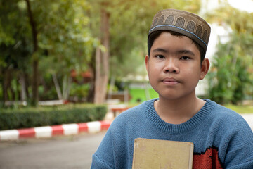 Portrait of asian muslim boy wears hat, sitting and holding a book on chest in the park area of his school, soft and selective focus, concept for lifestyle of boy around the world.