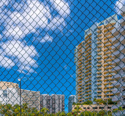 Modern apartments view behind chainlink fence in Miami, Florida. There is a chainlink fence at front with views of high-rise buildings with balconies against the sky with clouds at the background.