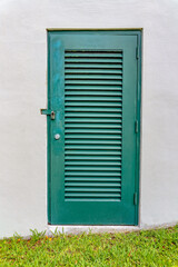 Fototapeta premium Green louvered door of a hidden electrical utility room of an irrigation at Miami, Florida. Door with padlock on a building with white wall and lawn outdoors.