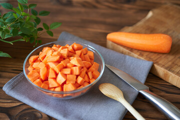 Cut carrots in a clear bowl on a gray cloth with a knife and wooden spoon. The concept is to prepare the carrots for cooking.