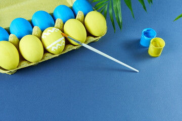 Multicolored Easter eggs packaged on a blue background with a brush and cans of paint. The concept is preparation for the holiday.