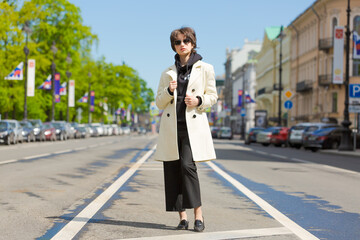 Hipster young girl with backpack enjoying in Central city, traveler in background city in a white coat and black sweatshirt in sunglasses young woman with a short haircut