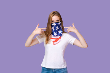 Young woman with mask in form flag america on her face in white t-shirt shows a gesture with her fingers at herself while standing on a purple background