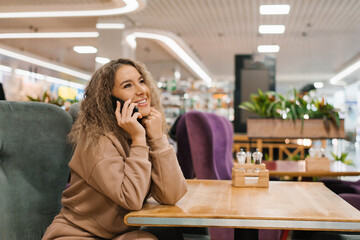 Young woman with curly hair is sitting at a cafe table and happily talking on a smartphone