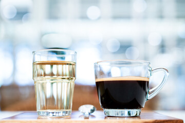 Crema of black coffee in a clear glass on a wooden tray, with a clear tea glass next to it selective focus, soft focus.           